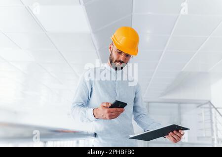 Using phone. Engineer in white clothes and orange protective hard hat standing and working indoors Stock Photo