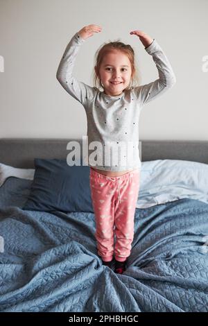 Little happy playful kid girl having fun making silly funny faces jumping on bed in bedroom in the morning at home Stock Photo