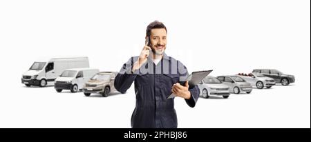 Male worker in a blue uniform using a mobile phone and holding a clipboard in front of parked vehicles isolated on white background Stock Photo