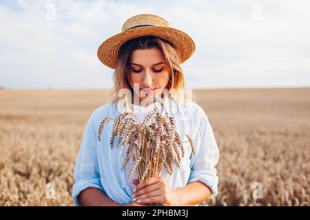 Portrait of young woman walking among wheat in summer field wearing straw hat holding bundle of ripe wheat at sunset. Stock Photo