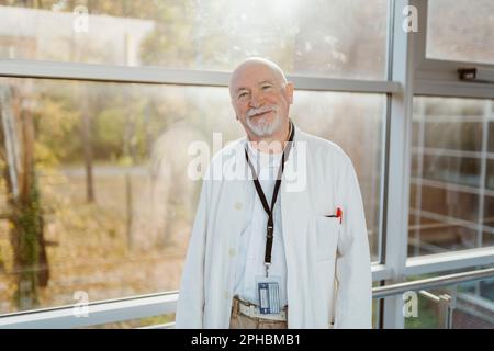 Portrait of smiling senior male doctor leaning on railing against window at hospital corridor Stock Photo