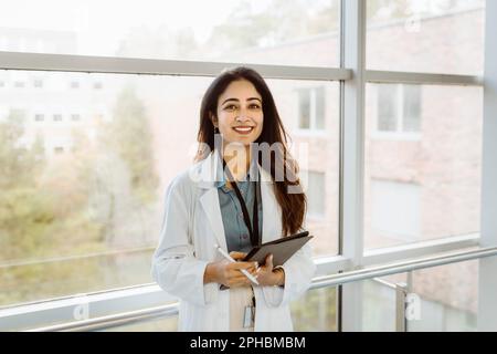 Portrait of smiling female doctor holding digital tablet and pen standing against window at hospital corridor Stock Photo