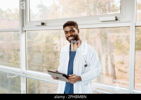 Portrait of smiling young male doctor holding digital tablet standing against window at hospital corridor Stock Photo