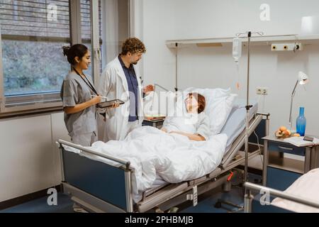 Healthcare workers talking to senior female patient lying on bed in hospital Stock Photo