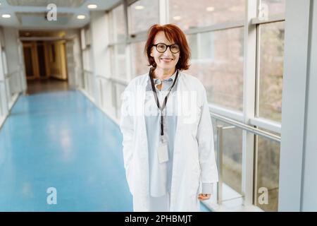 Portrait of smiling senior female doctor wearing ID card and lab coat standing in hospital corridor Stock Photo