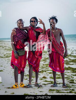 Three happy Tanzanian men in traditional clothing, standing together on a sandy beach, smiling towards the camera Stock Photo