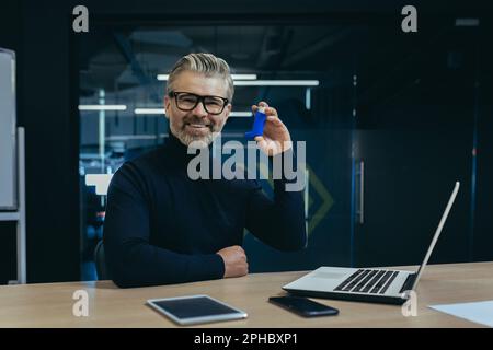 A gray-haired senior man sits at a desk in an office and holds an inhaler in his hand. Pointing and smiling at the camera. Stock Photo