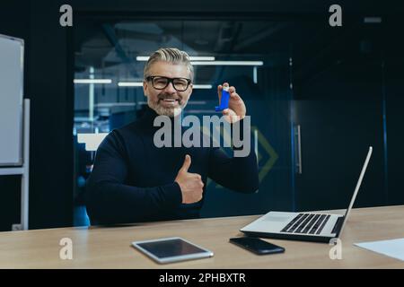 A gray-haired senior businessman sits at a desk in an office and holds an inhaler in his hand. Pointing and smiling at the camera. Super points, advertises, poses. Stock Photo