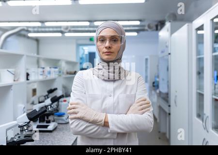Portrait of a young Muslim female lab technician, pharmacist in hijab and protective glasses standing in medical laboratory, arms crossed on chest, looking seriously at camera. Stock Photo