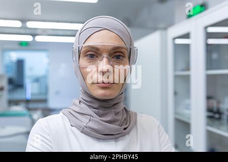 Close-up photo. Portrait of a young Muslim female lab assistant, pharmacist, scientist in hijab and protective glasses standing in medical laboratory, looking seriously at camera. Stock Photo