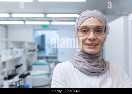 Close-up photo. Portrait of a young Muslim female lab assistant, pharmacist, scientist in hijab and protective glasses standing in medical laboratory, smilling, looking at camera. Stock Photo