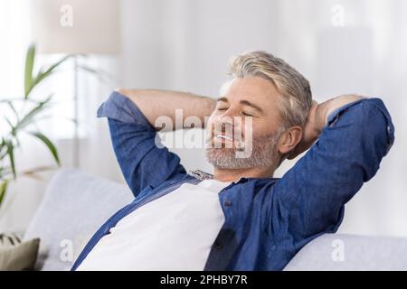Close-up photo. Senior gray-haired man sitting relaxed on sofa at home and resting, napping. Throwing his hands behind his head, he smiles. Stock Photo
