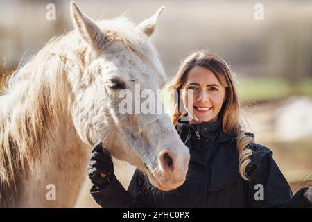 Young woman in black riding jacket standing near white Arabian horse smiling happy, closeup detail Stock Photo