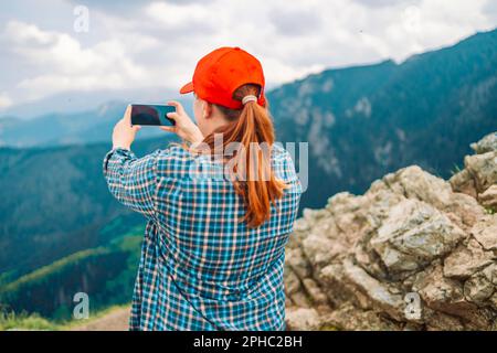 Young woman relaxing on the top mountains, holds a mobile phone in her hands and takes pictures of the landscape. Back view. Stock Photo