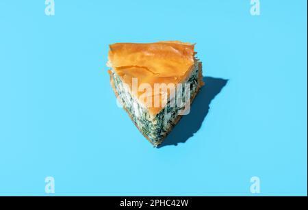 Single slice of phyllo cake, filled with spinach and feta cheese. Delicious spinach and cheese savory pie, in bright light on a blue table. Stock Photo