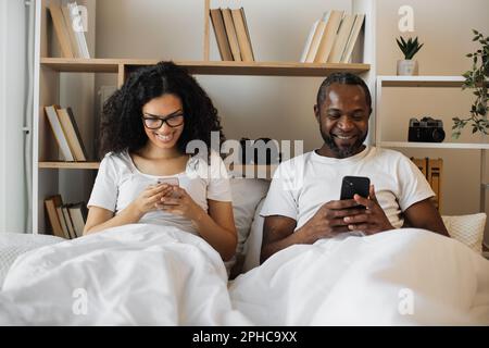 Serious multicultural husband and wife surfing internet using smartphones while sitting under cozy duvet in bedroom of modern flat. Busy spouses networking through social media via digital devices. Stock Photo