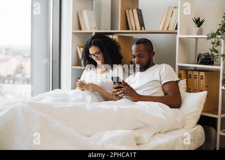 Serious multicultural husband and wife surfing internet using smartphones while sitting under cozy duvet in bedroom of modern flat. Busy spouses networking through social media via digital devices. Stock Photo