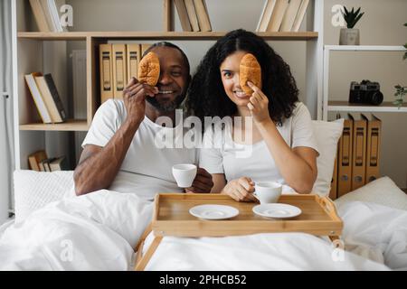 Cheerful multiethnic couple in love enjoying breakfast on bed tray early in morning at home. Smiling husband and wife having fun with croissants and coffee while spending weekend together indoors. Stock Photo