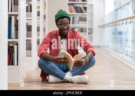 Happy African American millennial guy sitting on floor reading book while visiting library Stock Photo