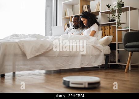 Lovely african american couple lying together in comfy bed and using modern laptop while robot vacuum cleaning wooden floor at home. Domestic chores with modern devices. Stock Photo