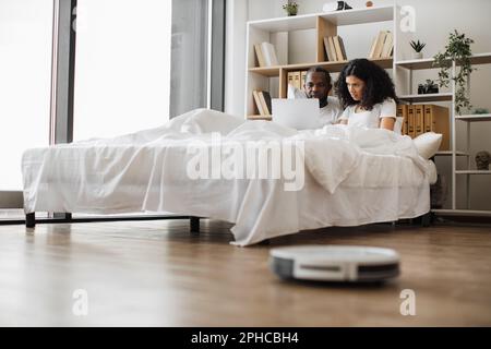 Multiracial family of two browsing internet on wireless laptop while resting together in comfy white bed. Modern vacuum cleaner removing dust from wooden floor on blurred foreground. Stock Photo