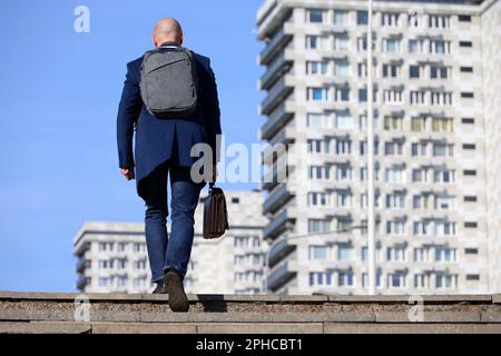 Businessman with backpack and leather briefcase in hand climbing stone stairs in city. Concept of career, success, moving to the top Stock Photo