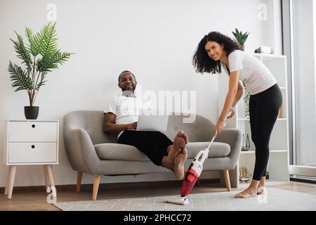 Multicultural woman vacuuming carpet at living room with cordless hand vacuum cleaner. Handsome husband resting on comfy couch with laptop on knees and smiling on camera. Family with gadgets at home. Stock Photo