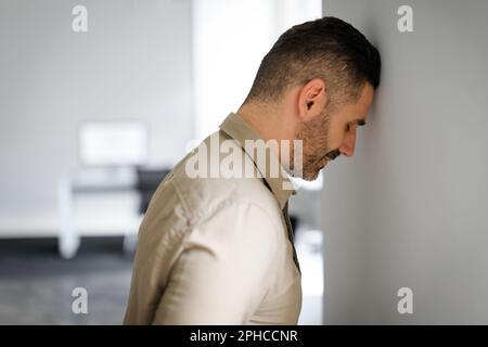 Upset middle aged businessman banging his head against wall in despair looking stressed, having problems at work Stock Photo