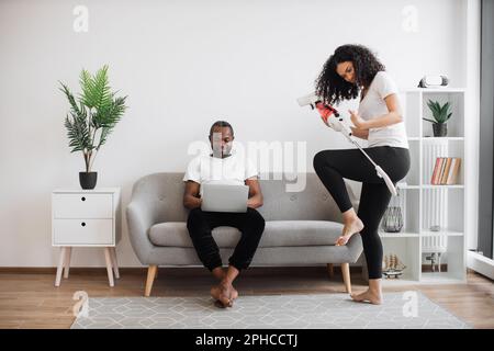 Cheerful african american woman in casual wear dancing with cordless vacuum cleaner during chores while focused husband sitting on couch and working on laptop. Lifestyles of modern young family. Stock Photo
