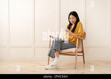 Happy asian lady sitting in armchair with smartphone, drinking coffee Stock Photo