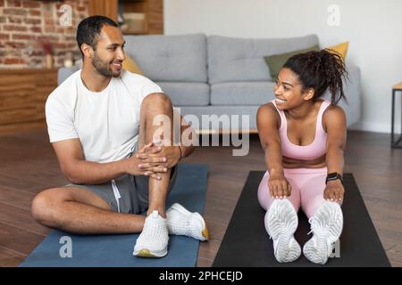 Glad young black couple in sportswear doing leg stretching exercises on mat, talking, rest, have fun Stock Photo