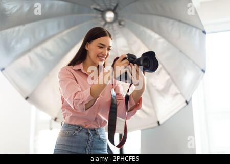Young female photographer standing in front of reflective umbrella, working with professional dslr camera in photostudio Stock Photo