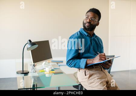 Portrait of thoughtful black man standing in office near workplace, holding clipboard and looking aside at free space Stock Photo