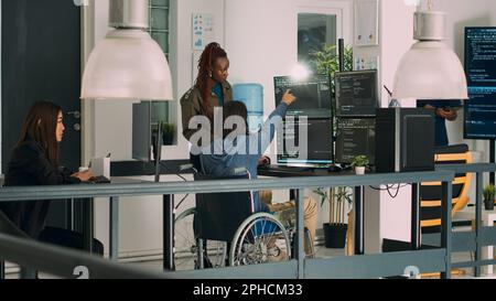Asian programer with chronic impairment working on software code with it engineer in agency office. App developer with disability in wheelchair using html script on temrinal window. Stock Photo