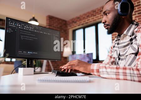 Database developer with wireless headphones programming algorithm using personal computer working remote in living room. African american freelance coder writing and debugging code at home. Stock Photo