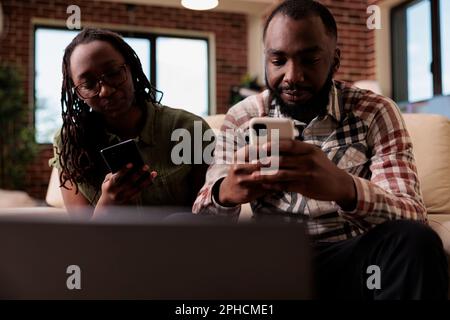 African american couple living together having issues not talking to each other spending time on smartphones sitting on couch. Boyfriend ignoring upset girlfriend after arguement in home living room. Stock Photo