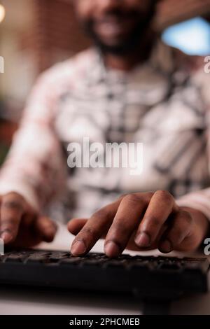 Closeup of african american coder writing software using computer keyboard while working remote from home living room. Selective focus on man freelancer hands typing text while programming. Stock Photo