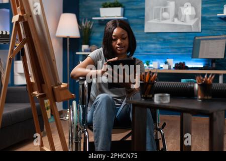 African american female art student wheelchair user taking remote art classes with tablet in home crafts studio. Young drawing schoolgirl using digital tablet to learn new sketching techniques. Stock Photo