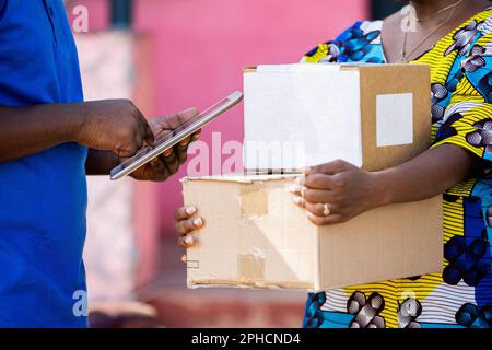 African Postal Delivery Man records receipt details of shipped cardboard box parcel with digital smart tablet bought online by female customer. Stock Photo