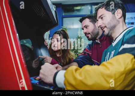 Caucasian friends in 90s attire play a vintage arcade game, two young men and a woman watch intently as one plays. Stock Photo