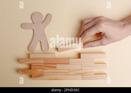 Woman putting wooden block to help human figure cross construction on beige background, top view Stock Photo