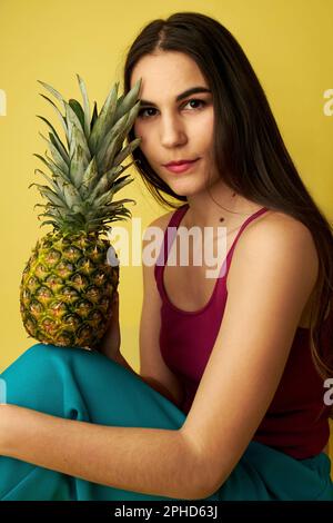 Woman holding a pineapple touching her face wearing a red shirt in front of a yellow background Stock Photo