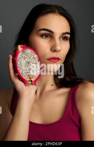 Woman holding half a cut dragon fruit up to face wearing a red shirt in front of a gray background Stock Photo