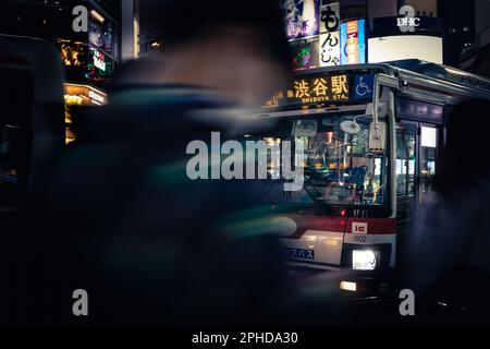 A young man uses his cell phone to navigate the streets while walking amidst the busy city nightlife of Tokyo as a Shibuya Station bus passes by. Stock Photo