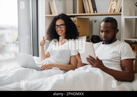 Pretty family woman keeping portable computer on blanket and showing thumb-up gesture while busy male texting on digital tablet. Multiracial couple in love lying in bed while working from home. Stock Photo