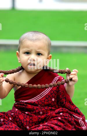 a cute bald baby boy is dressed up in Monk avatar, wearing a maroon shawl, Rudraksh, and giving a confused expression. Stock Photo