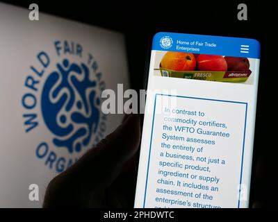 Person holding cellphone with webpage of World Fair Trade Organization (WFTO) on screen in front of logo. Focus on center of phone display. Stock Photo