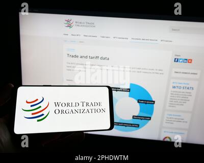 Person holding smartphone with logo of World Trade Organization (WTO) on screen in front of website. Focus on phone display. Stock Photo