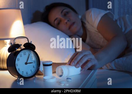 Mature woman taking bottle with pills in bedroom at night, focus on hand. Insomnia concept Stock Photo