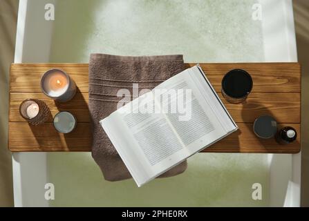 Wooden bath tray with open book, glass of wine and cosmetic products on tub indoors, top view Stock Photo